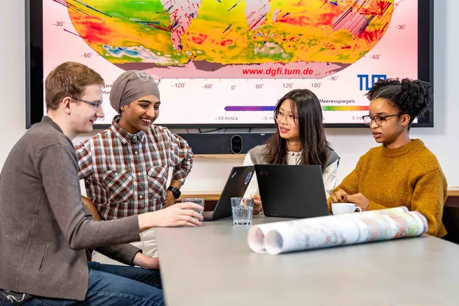 Vier Studierende sitzen am Tisch vor zwei Laptops und reden miteinander. Im Hintergrund ist eine Karte mit dem globalen Meeresspiegel zu sehen. 