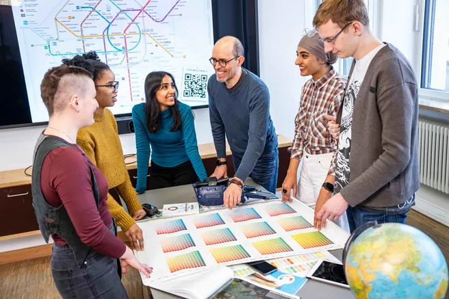 Researchers and students are standing at a table, on the table there are diverse maps and documents. In the background, there is a map of a metro network.