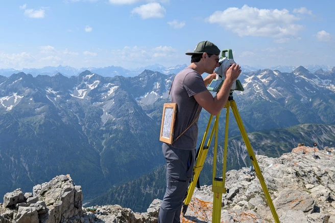 A geodesist stands at the top of a mountain and operates a measuring device.