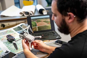A person is holding a drone in their hands. Next to them are a laptop and analog maps showing geodata and satellite images.